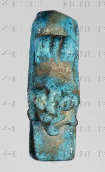Amulet — ram. Figurine of a ram lying on a flat base, with a loop for hanging on its back. Amulet; material unspecified. Sacred animal pendant; Late Period, Egypt, 664–332 BC. Height 2.9 cm; width 1.9 cm., A small three-dimensional object photographed against a plain light gray background, appearing to be a worn sculpted form with a textured surface. The object is predominantly bright turquoise blue with areas of darker teal and small black speckles, and it shows patches of brownish-beige deposits or residue on several raised and recessed areas. The shape has rounded protrusions and irregular bulges with a slightly glossy finish in places, and the surface shows fine irregular pits and roughness. The lighting produces soft highlights and subtle shadows that emphasize the object's contours and surface texture.