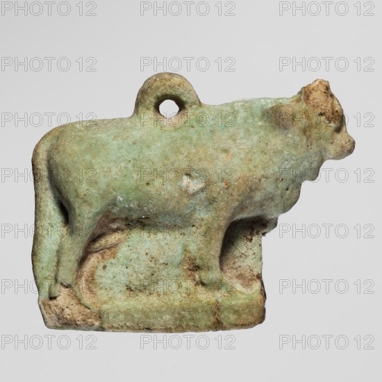 Amulet — bull; figurine of a bull standing on a base, full-plastic work with a pendant on the back; modeled quite carefully. Late Period, Egypt, 664–332 BC. Amulet; height 2.5 cm, width 3.1 cm., A small sculpted object in the shape of an animal is shown against a plain pale gray background; the object appears worn and has an overall greenish hue with areas of light blue-green and patches of beige and brown discoloration; the animal form includes a rounded body, a raised head with a blunt snout, a short tail, and four legs standing on a flat rectangular base, and there is a circular hole near the top of the body; surface texture looks rough and pitted with some smoother worn spots and mottled color variations.