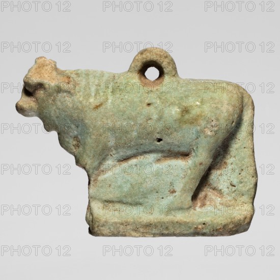Amulet — bull; figurine of a bull standing on a base, full-plastic work with a pendant on the back; modeling quite careful. Late Period, Egypt, 664–332 BC. Amulet; height 2.5 cm, width 3.1 cm., A small sculpted object in the shape of a standing animal, shown in profile facing left, with a loop or hole near its top; the surface shows a weathered, rough texture and uneven edges. The colors visible are pale green, mint, light turquoise, beige, tan, brown, and small areas of darker brown or rust. The form includes a raised base, a pronounced head with a short snout, a rounded body, and a short tail, with various pits and surface irregularities across the object.