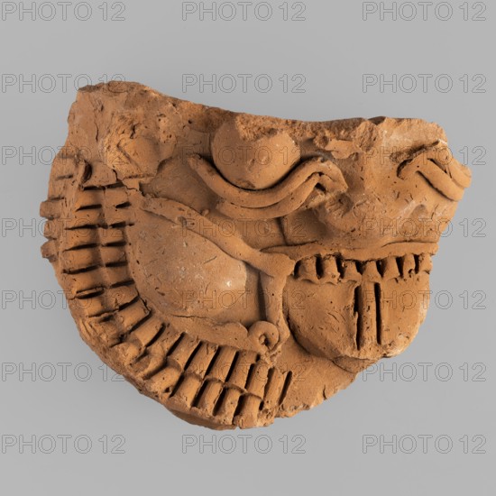 Mask of the god Bes — fragment of a vessel;, Bes myth., Egyptian deity; Egyptian art; 1st–2nd century AD Roman period. Found at Tell Atrib. Figurine; height 12.6 cm, width 9.5 cm., A broken fragment of a carved clay object in shades of terracotta and brown sits against a plain light gray background; the clay surface shows rough, textured areas and smoother rounded forms, with a curved raised band, a few rounded protrusions, and several linear grooves and incised marks forming repetitive ridged patterns and vertical slots, as well as irregular chipped edges and small pits.