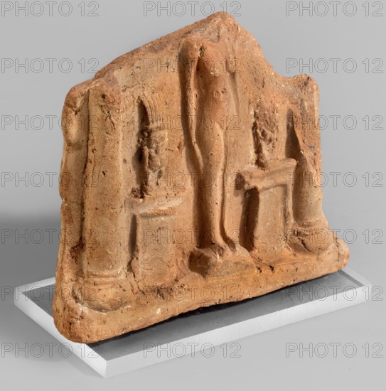 Fragment of a votive plaque in the form of a naos with the fertility goddess Isis-Hathor, and Bes;, Egyptian deities, Isis, naos, Bes; 664–30 BC, Late Period–Ptolemaic Period, Egypt; plaque; height 8.5 cm, width 10.4 cm, A weathered terracotta relief panel sits on a rectangular display base, showing a central standing human figure flanked by sculpted architectural elements and smaller seated or pedestal-mounted figures; the surface is rough with chips and pitting, and the colors visible are various shades of orange-brown for the terracotta, light gray for the display base, and neutral gray in the background.