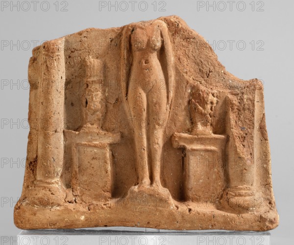 Fragment of a votive plaque in the form of a naos with the fertility goddess Isis-Hathor, and Bes, Egyptian deities, Isis, naos, Bes, 664–30 BC; Late Period–Ptolemaic Period, Egypt; plaque; height 8.5 cm, width 10.4 cm, The image shows a rectangular fragment of a terracotta relief in warm reddish-brown and sandy tones, featuring a central carved female figure standing upright on a raised base with elongated legs and arms held close to the body, flanked by two short pedestal-like supports topped by sculpted busts or heads, and bordered on each side by column-like vertical elements; the surface is textured with small pits and chips, and there are areas of lighter beige and darker brown discoloration across the worn, weathered material.