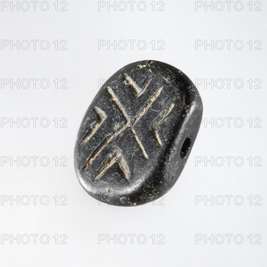Stamp seal with geometric ornament. A prism-shaped stamp seal with rounded edges and an oval, nearly rectangular base. A hole is drilled through the longer diagonal side walls transverse to the longitudinal axis of the stamp. On a flat base is carved a shallow geometric ornament consisting of regularly spaced, slightly open hooks: two large hooks with their tips touching in the center and four small hooks in triangular fields between the arms of the large hooks. Halaf culture, c. 6th–5th millennium BC. Northern Mesopotamia Iraq, Syria, Turkey. Seal. Height 0.8 cm; width 1.5 cm; depth 1.7 cm; hole diameter 0.2 cm., A small oval object with a smooth, worn surface in dark gray to black tones, showing lighter gray scratches or incisions forming a symmetrical pattern, with a visible rounded edge and a small drilled hole through one side, photographed against a pale, light gray background.