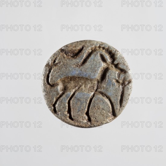 Conical stamp seal with a hole drilled below the top of the cone. On the circular base is engraved a walking gazelle shown in right profile, with a backward-curved notched horn, ear, eye, beard, and an upward-curving tail ending in a tassel of three engraved lines. Above the animal's rump is a horizontally displayed crescent moon; in front of the muzzle is a schematic plant or branch—vertical line ending in three short fan-like lines. Stamp seal depicting a goat gazelle, Northern Mesopotamian workshop, 9th–8th century BC previously dated ca. 9th–7th century BC. Origin: Northern Mesopotamia Iraq/Syria/Turkey. Seal; height 1.4 cm, width 1.65 cm., A circular, worn object with a raised, carved design depicting an animal figure and abstract shapes; the object shows a textured surface with rough edges and indentations, and the visible colors include gray, brown, and hints of blue.