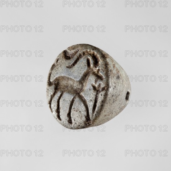 Conical stamp seal with a hole drilled below the top of the cone. On the circular base is engraved a walking gazelle shown in right profile with one backward-curved, notched horn, ear, eye, and beard. The upward-curving tail ends in a tassel of three engraved lines. Above the rump is a horizontally displayed crescent moon. In front of the muzzle is a schematic plant or branch with leaves depicted as a vertical line ending in three short fan-like lines. Materials: seal stamp seal. Style: Northern Mesopotamian. Date: 9th–8th century BC previously dated ca. 9th–7th century BC. Place of origin: Northern Mesopotamia Iraq/Syria/Turkey. Dimensions: height 1.4 cm; width 1.65 cm. Makership: unknown workshop. Subjects: goats/gazelle., A small rounded object with a worn, textured surface in shades of gray and brown, showing a carved outline of an animal with slender legs and a curved neck on one face, alongside a carved curved line and a small plantlike motif; the object has a drilled hole throug...