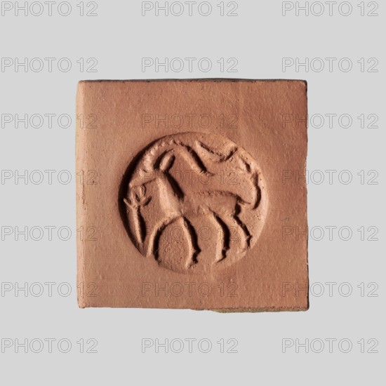 Conical stamp seal with a hole drilled below the top of the cone. On the circular base is engraved a walking gazelle shown in right profile: one backward-curved, notched horn, ear, eye, and beard. The upward-curving tail ends in a tassel of three engraved lines. Above the animal's rump is a horizontally displayed crescent moon. In front of the goat's muzzle is a schematic plant or branch—a vertical line ending in three short fan-like lines. Depicts a goat/gazelle. Northern Mesopotamian, 9th–8th century BC previously dated ca. 9th–7th century BC. Material: carved seal; height 1.4 cm, width 1.65 cm., A square terracotta-colored clay tile with a circular recessed carving in the center depicting a stylized animal shape; the tile surface is matte with subtle texture and small imperfections, edges are straight but slightly worn, and the visible colors are terracotta, reddish-brown, and a pale gray background.