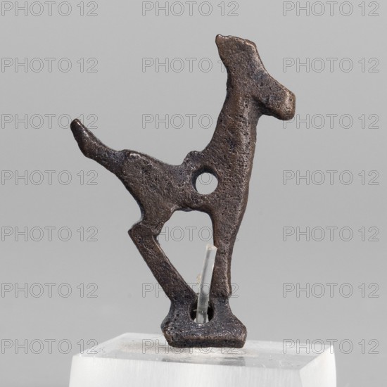 Figurine of a goat ibex, unknown Middle Eastern workshop; maker role/workshop: Semerau-Siemianowski, Wladyslaw 1849–1938 author. Miniature figurine of an ibex standing on a base in the form of a hollow prism, with single holes on the longer sides likely used to mount the figurine probably on a wooden handle. A round hole in the front of the ibex's torso may have served to attach ornaments e.g., bells., animal figurine, art of Iran, Eastern Gaulish scenography. Date: 1st half of the 1st millennium BC. Place of origin/discovery: Middle East; Asia Minor; Iran. Type: figurine. Dimensions: height 2.8 cm, width 2.1 cm, depth 0.6 cm., A dark brown, irregularly shaped metal object resembling an abstract animal silhouette is displayed against a light gray background, mounted on a transparent support that sits on a white rectangular base; the metal surface is rough and pitted with a circular hole through its midsection and a few thin clear plastic pins hold it in place.