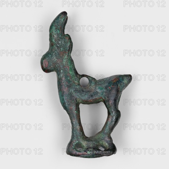 Figurine of an ibex goat standing on a hollow prism base with two pairs of holes on the longer sides, likely used to mount the figurine probably on a wooden handle. A round hole on the ibex's back may have served to attach ornaments e.g., bells. Terracotta or similar figurine; style: Eastern Near Eastern/Iranian. Date: 1st half of the 1st millennium BC. Provenance: Asia Minor / Iran, Middle East. Height 3.9 cm; width 2.5 cm; depth 1 cm., An irregularly shaped sculptural object resembling an abstract animal form, with a rough textured surface showing mottled green and dark green patina with areas of reddish-brown and darker gray, a hollow circular hole through its midsection, elongated neck or head feature curving upward, a rounded snout-like projection, a tapered rear or tail projection, slender leg-like supports creating an open space between them, and a flattened base with a couple of small voids, all set against a plain light gray background.