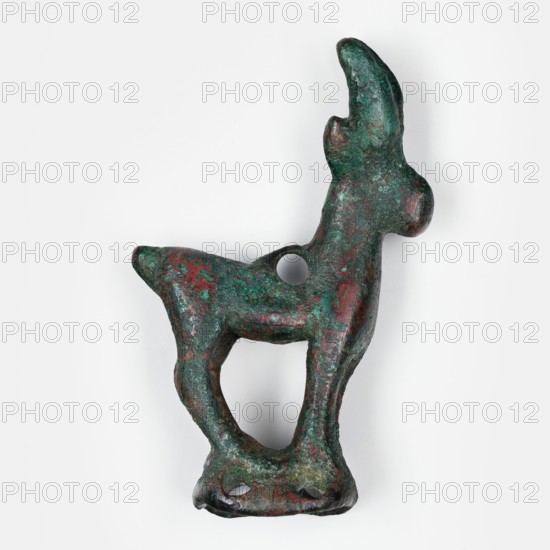 Figurine of an ibex standing on a base in the form of a hollow prism, with two pairs of holes on the longer sides used to mount the figurine probably on a wooden handle. A round hole on the ibex's back may have served to attach ornaments e.g., bells. Terracotta/unknown material, animal figurine; ibex. 1st half of the 1st millennium BC. Asia Minor / Iran Middle East. Height 3.9 cm, width 2.5 cm, depth 1 cm., A small sculptural object shaped like an animal with upright ears and a rounded snout, shown against a plain white background; the object has a roughly textured surface with a mottled patina in green and dark green with areas of reddish-brown and traces of darker brown, featuring a rounded hole through the body and an open space beneath formed by extended legs and a base with two small openings.