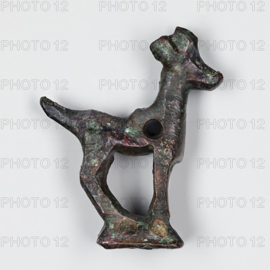 Figurine of a goat, unknown Near Eastern workshop; ibex, animal figurine, metalwork. Figurine of a goat standing on a base in the form of a hollow prism, with two pairs of holes on the longer sides, used to mount the figurine probably on a wooden handle. A round hole is also located in the front of the ibex's torso and may have been used to attach ornaments, e.g., bells. One of two similar goats. 1st half of 1000 BC. Asia Minor, Iran. Figurine; height 3.8 cm, width 2.8 cm, depth 1 cm., A small sculptural object shaped like an animal with a raised head and a pointed tail, showing a rough, uneven surface with visible patina in green and brown tones and touches of dark reddish and black; the form has an open triangular space between its slender legs, a round hole in the body, and a flat base with two rounded protrusions, all photographed against a neutral light-gray background.