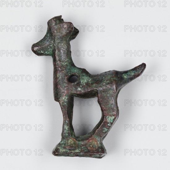 Figurine of a goat, unknown Near Eastern workshop. Ibex animal figurine in metals/metalwork. Goat standing on a base in the form of a hollow prism with two pairs of holes on the longer sides, likely used to mount the figurine probably on a wooden handle. A round hole in the front of the ibex's torso may have been used to attach ornaments e.g., bells. One of two similar goats. 1st half of 1000 BC. Origin: Asia Minor/Iran. Figurine. Height 3.8 cm; width 2.8 cm; depth 1 cm., A small sculptural object shaped like a stylized animal with a raised head, elongated neck, rectangular body, and tapered tail, showing a rough textured surface with patches of green and dark brown patina, a round hole through the body, an open arch between the legs, and a flat base with slight irregularities.