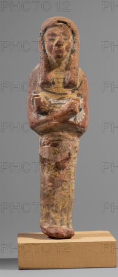 Ushabti, Restitution. Hieroglyphs; funerary figurine; wig; hoes; grave goods; mummy-shaped figurine. Arms crossed on the chest, right folded over left, holding hoes in each hand. Rectangular basket slung over both shoulders on the back. Painted tools. Wide painted usekh necklace on the chest; bracelets on the wrists. Long three-part wig, ears uncovered. Round face with large eyes, wide nose, mouth barely visible; wig and facial details painted. Painted inscription around the figurine's legs, mostly erased; text illegible. Circa 1295–1186 BC; New Kingdom, 19th Dynasty, Egypt. Shabti. Height 15.1 cm; width 4.2 cm., A weathered small statue of a human figure with crossed arms, standing on a rectangular base, showing a rounded head and a stylized wig or hair that frames the face; the surface has worn areas and textured pitting. The visible colors include reddish-brown, muted yellow, pale beige, black accents, and traces of white, with the base in a tan or light brown hue. The figure's tors...