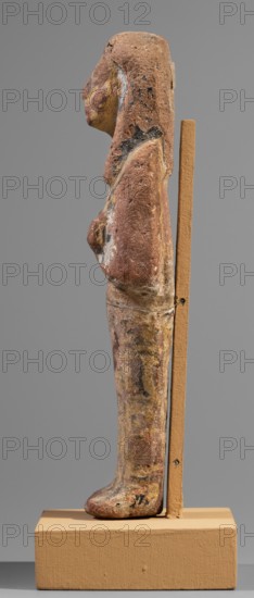 Ushabti, Hieroglyphs; funerary figurine; mummy-shaped shabti, c. 1295–1186 BC, New Kingdom, 19th Dynasty, Egypt. Height 15.1 cm; width 4.2 cm. Arms crossed on the chest, right folded over left, holding hoes in each hand. Rectangular basket slung over both shoulders on the back. Painted tools. Wide painted usekh necklace on the chest; bracelets on the wrists. Long three-part wig, ears uncovered. Round face with large eyes, wide nose, mouth barely visible; wig and facial details painted. Painted inscription around the legs, mostly erased and illegible., A weathered vertical figurine mounted on a rectangular base, shown in profile facing left, with a carved head, torso, and legs; the surface is rough and pitted with worn paint in shades of reddish-brown, yellow, black, white, and patches of pale tan, and the figure leans against a narrow upright support of a tan color attached to the base; the head has a smoothed contour with traces of pigment, the torso shows a raised chest area and a na...