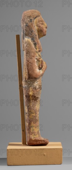 Ushabti, Restitution. Hieroglyphs; funerary figurine; wig; hoes; grave goods; mummy-shaped figurine. Arms crossed on the chest, right folded over left, holding hoes in each hand. Rectangular basket slung over both shoulders on the back; painted tools. Wide painted usekh necklace on the chest; bracelets on the wrists. Long three-part wig, ears uncovered. Round face with large eyes, wide nose, mouth barely visible; wig and facial details painted. Painted inscription around the legs, mostly erased and illegible. c. 1295–c. 1186 BC; New Kingdom, 19th Dynasty, Egypt. Shabti. Height 15.1 cm; width 4.2 cm., A weathered vertical figurine mounted on a rectangular base is shown in profile, supported by a thin vertical rod behind it; the figurine has a rounded head and a long, slender body with a pronounced chest and a single visible foot at the bottom. The surface of the figurine appears rough and uneven with visible flaking and patches of paint in earthy tones including brown, tan, yellow, ochr...
