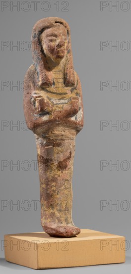 Ushabti, Hieroglyphs; funerary figurine shabti, mummy-shaped figurine, c. 1295–1186 BC, New Kingdom, 19th Dynasty, Egypt. Height 15.1 cm; width 4.2 cm. Arms crossed on the chest, right over left; hoes in each hand. Rectangular basket slung over both shoulders on the back. Painted tools. Wide painted usekh necklace on the chest; bracelets on the wrists. Long three-part wig, ears uncovered. Round face with large eyes, wide nose, mouth barely visible; wig and facial details painted. Painted inscription placed around the figurine's legs, mostly erased and illegible., A small weathered figurine stands on a rectangular base, showing a humanoid form with a stylized head and a long narrow body; the head has a rounded headdress framing a worn face, and the upper torso displays folded arms across the chest. The surface appears rough and pitted with flaking paint, and the visible colors include muted reds, faded yellows, pale whites, dark gray or black accents, and areas of brownish and beige ton...