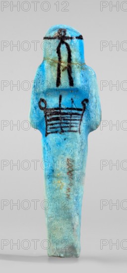 Amenhotep's shabti with baskets on the back; arms crossed on the chest, right folded over left, holding hoes in both hands; rectangular basket with loops on the back; painted tools. Three-part long smooth wig with headband covering ears; face damaged; painted headband. Painted inscription in one column on front of legs reading Osiris the father of God ... scribe of the temple of Amun-Re, Amenhotep. Ca. 1069–715 BC, Third Intermediate Period, 21st–23rd Dynasty, Egypt. Shabti, height 13.3 cm, width 4.2 cm., A small figurine-shaped object stands upright against a neutral background, predominantly bright turquoise and light blue with areas of pale green and hints of darker blue; a few black painted linear motifs appear on the upper body and chest, and there are traces of brownish discoloration and tiny flecks of gold on the surface; the form has rounded shoulders, a bulbous head, and an elongated narrow lower body with a slightly irregular, textured surface.