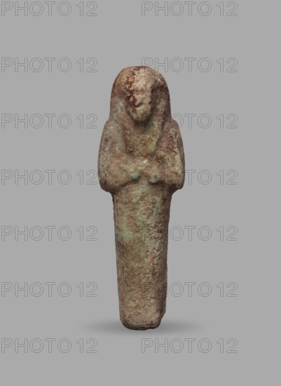 Ushabti, Mummy-shaped funerary figurine, ushabti. Arms crossed on the chest, hands arranged in a line next to each other, holding hoes in both hands with slightly convex tools. Wears a long, three-part smooth wig with ears uncovered. Round face with illegible facial details. Not inscribed. Ca. 1069–715 BC; Third Intermediate Period, 21st–23rd Dynasty, Egypt. Height 5.5 cm; width 1.8 cm., A weathered vertical statuette with a rounded top and an elongated lower section standing against a plain background, showing a rough, pitted surface texture; the upper portion suggests a simplified head and shoulders with shallow, smoothed facial features and a suggestion of crossed arms at chest level; the surface displays mottled tones of brown, tan, green, and gray with areas of darker brown patina and lighter greenish corrosion, and a soft shadow beneath the base.