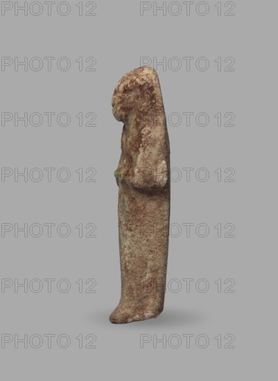Ushabti, Mummy-shaped funerary figurine, ushabti. Arms crossed on the chest, hands arranged in a line next to each other, holding hoes in both hands with slightly convex tools. Long, three-part smooth wig leaving ears uncovered. Round face with illegible facial details. Not inscribed. ca. 1069–715 BC; Third Intermediate Period, 21st–23rd Dynasty, Egypt. Height 5.5 cm; width 1.8 cm., A small vertical statue of a standing figure shown in profile against a plain gray background, with a textured surface in shades of brown, tan, and hints of green; the figure has a rounded head and a long, narrow body with subtle raised areas suggesting folded arms and a gently protruding foot, the surface appearing weathered and mottled with lighter and darker brown patches and small greenish spots.