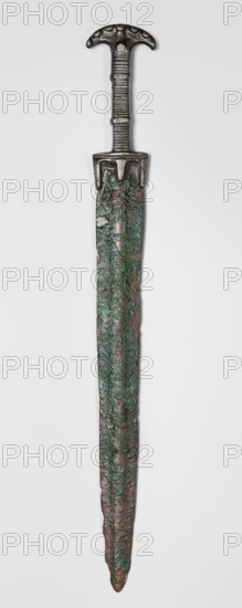 Double-edged sword, Bosporan workshop. Short sword, cast in one piece. Double-edged blade with a centric point and a prominent bulge running through the center of both faces. Hilt divided into three segments: a crescent-shaped pommel, decorated on both sides with small oval knobs imitating rivet heads; one such knob at the transition to the hilt shaft on each side. b square-section shaft decorated on two opposite sides with horizontal incisions; shaft is encircled in two places by a convex, flat band; guard at base of blade. c guard in the form of a horizontal profiled strip from which three protrusions emerge — two longer along the blade line and one shorter on the axis extension. 6th century BC, Black Sea region Bosporan, weapon. Height 45.3 cm; width 5.6 cm; depth 1.7 cm; length 45.5 cm; weight 0.566 kg., An elongated metal object with a dark metallic handle and a rounded, T-shaped pommel showing patterned indentations, a ridged grip, and a short guard; below the guard a long narrow...