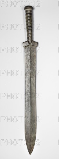 Double-edged sword, Bosporan workshop. Militaria, melee weapon from the Black Sea region; short sword cast in one piece. Double-edged blade with a centric point. A slight protrusion, called the axis, runs through the center of both flats. The handle is divided into three parts: a the pommel in the form of a small, slightly flattened mushroom cap; b the handle shaft with round cross-section, narrowing towards the blade, formed from 9 rings separated by grooves; c the guard in the form of oblique short leaves protruding slightly beyond the width of the blade and separated by a triangular bulge. Sword of the Cimmerian type, 6th century BC, unknown origin Black Sea region. Weapon dimensions: height 55.5 cm, width 5.8 cm, depth 3.5 cm, weight 0.888 kg, length 55.3 cm., A long metal sword viewed vertically against a light background, showing a rounded pommel and a ribbed handle with a short crossguard, the blade broad and tapering to a rounded point; visible colors include gray, dark gray, g...