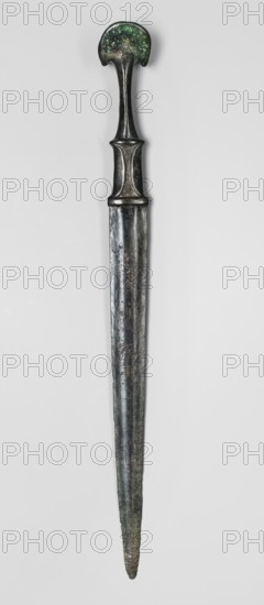Dagger, unknown Luristan workshop. A fully cast Luristan bronze dagger with a full hilt and a flared pommel. The pommel, shaped like two angled, opposing crescent-shaped lobes, merges into a narrow shaft that continues into a rectangular element imitating a casing with curved mounting plates at the bottom of the hilt such hilts also had casings of various materials applied. The hilt merges directly into a double-edged blade that tapers evenly toward the tip. A ridge runs from the tip of the engraving toward the center of the superstructure, which after a short distance splits, creating a long, narrow, triangular flat field. 9th–6th century BC; Iron Period II. Luristan, Iran. Weapon. Height 35.7 cm; width 3.2 cm; depth 3.4 cm; weight 264 g., A long metallic object with a rounded top that has a mottled green and dark patina, a narrow curved neck leading to a wider midsection with subtle raised ridges, and a tapering blade-like lower portion showing a dark silvery and greenish surface wit...