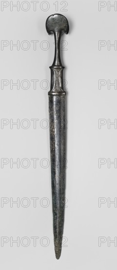 Dagger, unknown Luristan workshop. A fully cast dagger with a full hilt and a flared pommel. The pommel, shaped like two angled, opposing crescent-shaped lobes, merges into a narrow shaft, which merges into a rectangular element imitating a casing with curved mounting plates at the bottom of the hilt such hilts also had casings of various materials applied. The hilt merges directly into a double-edged blade, which tapers evenly towards the tip. A ridge runs from the tip of the engraving towards the centre of the superstructure, which after a short distance splits, creating a long, narrow, triangular flat field. Luristan bronzes; daggers; bladed weapons; militaria; material culture. 9th–6th century BC; Iron Period II, Luristan, Iran. Weapon. Height 35.7 cm; width 3.2 cm; depth 3.4 cm; weight 264 g., A long metallic object with a dark gray to black patina, showing worn and slightly rough surfaces, photographed against a pale gray background; the object tapers to a pointed end and has a w...