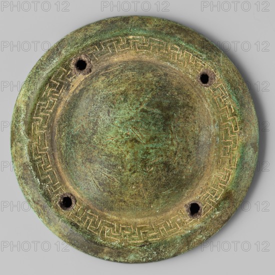 Shield boss, Bosporan workshop; round shield boss with a regularly convex central part, surrounded by a narrow zone decorated with an incised meander ornament and four symmetrically arranged round holes; another narrow undecorated zone set diagonally, flowing into a low vertical edge; shield boss, weight 0.166 kg; 6th–5th century BC; Black Sea areas., A round, shallow metal object with a raised central dome and a wide rim, showing a patina of green and brown with areas of darker brown and hints of turquoise; the rim bears a repeated geometric band in a lighter brass color, and there are a few irregular holes near the rim with dark interiors; the surface has subtle scratches and uneven texture, with mottled patches of green and brown across the dome and rim, all set against a neutral pale gray background.