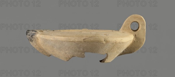 Oil lamp with an image of Helios. Roman single-burner oil lamp made from a mold. Round, flat container with smooth, flat arms; one arm has an ingot. Vertical vice opposite the burner decorated with two grooves. Round, slightly concave discus bounded by three concentric grooves; center shows a bust of Helios. Next to the bust is a round olive hole made after the lamp was pressed from the mold. Burner short, incorporated into the arms, round Bussière type 2b, blackened. Bottom and part of the burner are broken. Light clay may indicate manufacture in Corinth Bernhard suggests an eastern workshop. 1st century., A broken shallow bowl or lamp-shaped ceramic object in beige and tan tones with a rounded rim and a curved hooked handle on one side that has a circular hole, a darker worn tip on the opposite edge showing black and gray discoloration, rough textured surfaces with chips and missing fragments along the lower edge, and a neutral gray background.