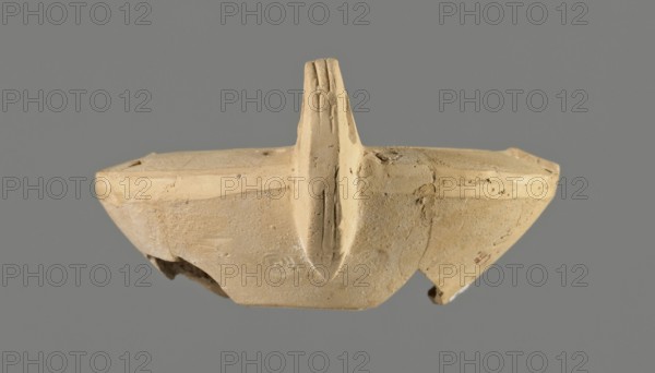 Oil lamp with an image of Helios, unknown Greek workshop, Single-burner oil lamp, made from a mold. Round, flat container with smooth, flat arms; one arm has a so-called ingot. Vertical vice opposite the burner decorated with two grooves. Round, slightly concave discus bounded by three concentric grooves; center shows a bust of Helios. Next to the bust is a round olive hole made after the lamp was pressed from the mold. Short, incorporated round burner Bussière type 2b, blackened. Bottom and part of the burner are broken. Light clay may indicate original manufacture in Corinth Bernhard suggests an Eastern workshop. 1st century., A broken beige clay object with a central raised fin-like ridge is shown against a solid gray background; the surface of the clay appears rough and worn with small chips and cracks, and there are a few darker spots and smudges on the beige surface, with subtle variations of light beige and tan tones across the piece.