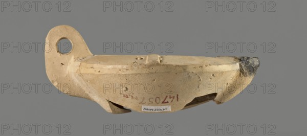 Oil lamp with an image of Helios. Single-burner Roman oil lamp, Broneer Lamp type XXVII C, made from a mold. Round, flat container with smooth, flat arms; one arm has an ingot. Vertical vice opposite the burner decorated with two grooves. Round, slightly concave discus bounded by three concentric grooves; center bears a bust of Helios. Next to the bust is a round olive hole made after molding. Burner short, incorporated into the arms, round Bussière type 2b, blackened. Bottom and part of the burner are broken. Light clay may indicate production in Corinth Bernhard suggested an Eastern workshop. 1st century; oil lamp., A small beige clay vessel with a rounded body and a raised rim, showing rough and pitted surface texture and a few chips and cracks; one end has a flat, ear-like extension with a circular hole, and the opposite rim end is darker with soot-like dark gray discoloration; the vessel rests against a neutral gray background and displays subtle variations of beige, tan, light br...