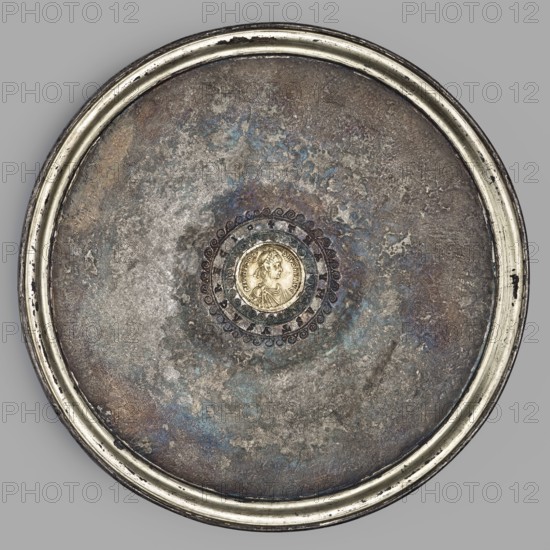 Platter with a gold medallion of Theodosius II. Flat silver platter on a ring-shaped foot with profiled inner edges. In the central part is a coin/medallion of Theodosius II reigned 408–450. Made after 430, Roman/Byzantine Empire. Cast using a foundry mold; foot cast separately and soldered on. The coin was set into a precisely fitted hole. Gilding on the frame and a stripe along the rim applied by amalgam chemical gilding. Inscription letters struck with single stamps. Ornament punched; spiral elements joined by brazing. Traces of filing/smoothing visible on the underside of the bowl from manufacture. Material: silver with gold medallion; foundry, casting and punching techniques. Vessel; diameter 18.6 cm, height 2 cm, weight 543.71 g., A circular metal object with a raised rim in a light silver color surrounds a weathered central surface showing mottled tones of gray, brown, blue, and hints of green and purple; the center contains a round, slightly raised medallion in a warm gold colo...