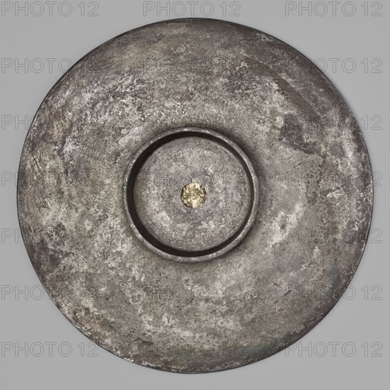 Platter with a gold medallion of Theodosius II. Flat silver platter on a ring-shaped foot with profiled inner edges; in the central part is a gold coin/medallion of Theodosius II reigned 408–450. Made after 430 CE. Cast using a mold of foundry earth; the foot was cast separately and later soldered on. The coin was fitted precisely into a purposely made hole. Gilding on the frame and a stripe along the rim was likely done by amalgam chemical gilding. Inscription letters were struck with single stamps. Ornament executed by punching; spiral elements joined using bending. Traces of filing smoothing visible on the underside of the bowl related to manufacture. Byzantine/Roman Empire workmanship. Vessel; diameter 18.6 cm, height 2 cm, weight 543.71 g., A large circular metallic object fills the frame, showing a textured surface with mottled shades of gray and silver and hints of darker charcoal and muted brown; a recessed central ring creates a raised inner disk with similar rough texture, an...