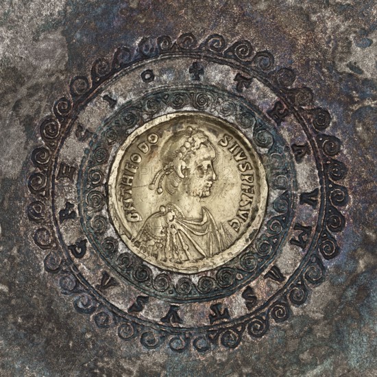 Platter with a gold medallion of Theodosius II, Byzantine workshop. Flat platter on a ring-shaped foot with profiled inner edges. In the central part is a coin of Theodosius II 408–450. Made by casting, probably in a mold of foundry earth; the foot was cast separately and later soldered on. The platter was likely made with a specific coin in mind, as shown by the precisely fitted hole. Gilding on the frame was most likely done using the amalgam chemical gilding technique; parts of the bowl, such as the stripe along the rim, were also gilded this way. The letters of the inscription were struck with single stamps. Ornamentation was applied with punching; spiral elements are additionally joined using boning. Traces of filing smoothing related to the manufacturing technique are visible on the underside of the bowl. After 430, Byzantine/Roman Empire. Vessel; silver with gold; medallion; diameter 18.6 cm; height 2 cm; weight 543.71 g., A circular metallic object with a raised central medalli...