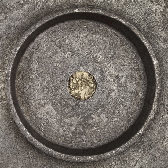 Platter with a gold medallion of Theodosius II, Byzantine workshop. Flat platter on a ring-shaped foot with profiled inner edges. In the central part of the platter is a coin of Theodosius II 408–450. Made using casting, probably in a mold of foundry earth; the foot was cast separately and later soldered on. The platter was likely made to fit a specific coin, as shown by the precisely fitted hole. Gilding on the frame and a stripe along the rim was likely done by the amalgam chemical gilding technique. Inscription letters were struck with single stamps. Ornament executed by punching; spiral elements additionally joined using boning. Traces of filing smoothing of the surface are visible on the underside of the bowl. After 430, Roman Empire 27 BC–476. Found in Toulouse. Vessel; diameter 18.6 cm, height 2 cm, weight 543.71 g., A circular metal surface with a raised rim surrounds a textured interior in shades of gray and silver, with a small round golden area at the center that shows a few...