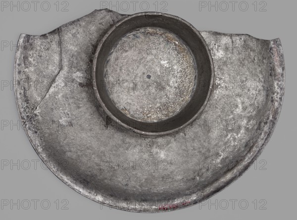 Platter with a depiction of a boar, Byzantine workshop, boar iconography, platters, 2nd half of the 5th century AD, Toulouse, Roman Empire 27 BC–476, vessel, weight 505.81 g, A worn round metal object with a raised circular rim sits against a plain background, showing a mottled surface texture that appears rough and pitted. The object displays various shades of gray with areas of darker gray and lighter silvery highlights, plus small patches of brownish discoloration and faint hints of rust-like tones. The rim and inner depression have darker, shadowed gray, while the flatter outer area shows lighter, uneven gray and silvery wear. The overall shape is circular with two small flat extensions at the top edges, and the surface shows scratches, scuffs, and irregular speckled patterns.