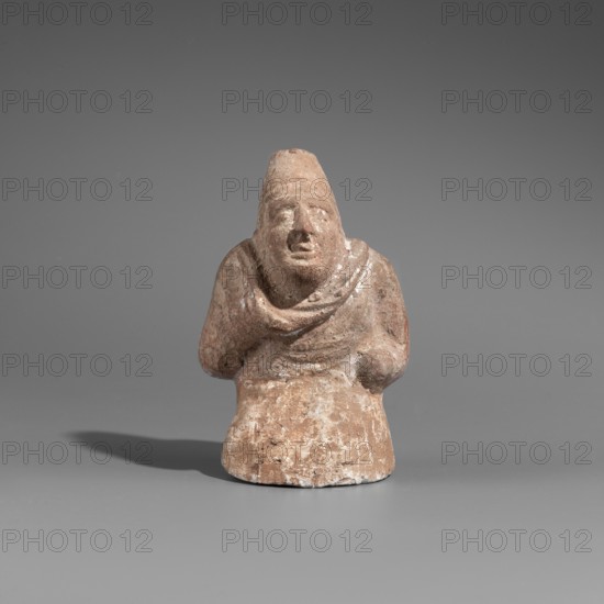 Hunchback puppet. Figurine hollow inside. Hunchbacked torso with a large head in a pointed cap set on a short neck. Elongated face with a large hooked nose and full-lipped mouth. Large bulging eyes with traces of white paint on the pupils. Torso wrapped in a cloak forming a thick fold around the neck and covering the right arm raised toward the chin. Left arm bent, resting at the waist. Opening on the side under the elbows for attaching the legs. Opening at the top of the cap for attaching strings. Traces of polychrome with white paint on the torso. 1st century BC, Hellenistic period. Myrmekion findspot; Pantikapaion origin. Figurine. Height 8.6 cm; width 5.5 cm; depth 4.6 cm., A small, worn terracotta figurine in shades of brown and beige sits against a smooth gray background, showing a rounded base and a hooded or conical head with a faintly detailed face; the figure's shoulders are draped in a sculpted shawl or cloak and its arms rest close to the torso, with one hand slightly raise...