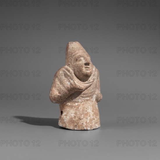 Hunchback puppet, Figurine hollow inside. Hunchbacked torso with a large head wearing a pointed cap set on a short neck. Elongated face with a large hooked nose and full-lipped mouth. Large bulging eyes with visible traces of white paint on the pupils. Torso wrapped in a cloak forming a thick fold around the neck and covering the right arm raised toward the chin. Left arm bent, resting at the waist. Opening at the sides under the elbows for attaching the legs. Opening at the top of the cap for attaching strings. Traces of polychrome with white paint on the torso. 1st century BC, Hellenistic period. Found at Myrmekion; origin Pantikapaion. Figurine; height 8.6 cm, width 5.5 cm, depth 4.6 cm., A small weathered terracotta figure stands against a smooth gray background, showing a rounded head with a pointed cap and a broad, simplified face with a pronounced nose and slightly open mouth; the figure’s torso is wrapped in draped folds suggesting a cloak, with one arm bent across the chest an...