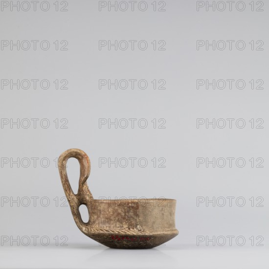 Miniature kyatos, A miniature kyatos with a flat bottom and a bowl whose walls widen diagonally almost halfway up and then become vertical. Where the walls break, a sharp protrusion remains on the outer side the diameter of the upper part is smaller than the lower. The vertical vise is strongly extended upwards, attached to a protrusion on the body and to the spout, with a crossbar above the spout's edge. On the outer side it has a ribbon shape, then from the top widens inward with an undercut above the spout. The undercut part is decorated with engraved vertical lines. Engraved lines are also found on the bottom arranged in a grid and under the base of the vise in the shape of the letter X. The upper surface of the edge on the body is decorated with diagonal engravings, the lower part of the vertical walls with recessed dots. Both rows are separated by a horizontal engraved line. Early 7th century BC, Caere; vessel, height 6.3 cm, width 7.7 cm., A small shallow ceramic vessel sits on ...