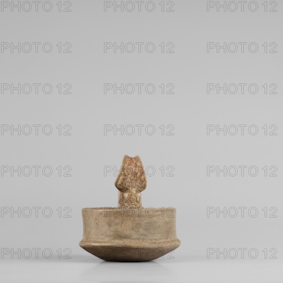A miniature kyatos with a flat bottom and a bowl whose walls widen diagonally almost halfway up and then become vertical. Where the walls break, a sharp protrusion is left on the outer side the diameter of the upper part is smaller than the lower. The vertical handle is strongly extended upwards, attached to a protrusion on the body and to the spout, with a crossbar above the spout's edge. On the outer side the handle has a ribbon shape, then from the top widens inward with an undercut above the spout. The undercut part is decorated with engraved vertical lines. Engraved lines are also found on the bottom arranged in a grid and under the base of the handle in the shape of the letter X. The upper surface of the rim on the body is decorated with diagonal engravings, the lower part of the vertical walls with recessed dots. Both rows are separated by a horizontal engraved line. Early 7th century BC, Caere; vessel; height 6.3 cm, width 7.7 cm., A small sculpted object sits centered on a pla...