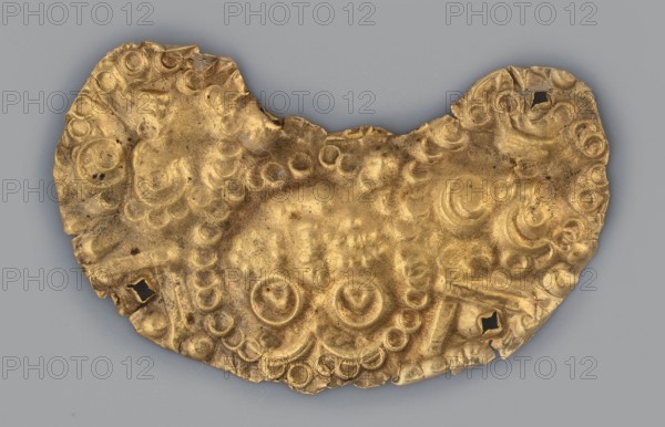 Crescent-shaped appliqué with three heads; appliqué made of thin gold sheet in the shape of a moon, decorated with a depiction of three stylized heads. The central, largest, has triangular, convex pupils surrounded by a circle. The two side eyes consist of two concentric circles and a marked mouth formed by two small, horizontal, convex lines. Each face is surrounded by a series of small circles. The appliqué has four perforations for sewing the plate, e.g. to a plaque. 5th–4th century BC, Greece . Gold; appliqué; height 2 cm, width 3 cm, depth 0.1 cm, weight 1 g., A curved, irregularly shaped metallic object with a golden color rests against a gray background, showing a textured surface of raised circular and spiral motifs and depressions; the edges are uneven and slightly torn in places with a few small square holes near the rim, and the surface displays areas of darker tarnish and subtle variations in gold tone.