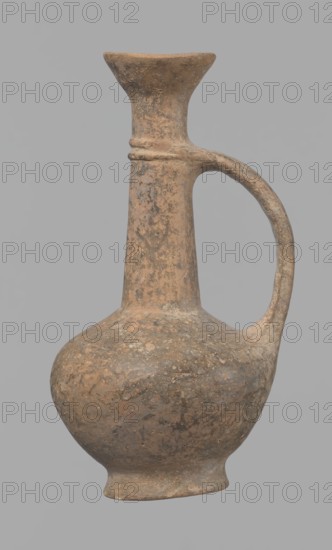 Jug, Cypriot pottery, Base Ring Ware I. Jug with an echinus-shaped foot; a biconical, slightly rounded belly; a high narrow neck terminating in a funnel-shaped, flared spout. The neck is set at a slight inclination from the plane of the foot. A ribbon-shaped handle is mounted two-thirds up the neck and attached to the body; at the neck it merges into a double applied ring surrounding the neck. Pinkish clay with a brown surface, in places fading to a brick-red hue. Surface heavily rubbed with slight loss of the rings on the neck. 1600–1450 BC, Cyprus. Vessel; height 12.3 cm, diameter 6.7 cm., A worn, rounded ceramic vessel with a narrow neck and a flared rim, featuring a single curved handle connecting the neck and shoulder; the surface shows a mottled, rough texture with patches of earthy brown and muted tan, along with areas of darker brown and gray; the background is a uniform light gray.