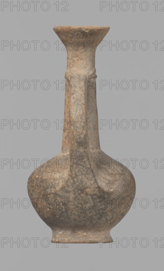Jug, Cypriot pottery, Base Ring Ware I, 1600–1450 BC. Jug with an echinus-shaped foot; a biconical, slightly rounded belly; a high, narrow neck terminating in a funnel-shaped, flared spout. The neck is set at a slight inclination from the plane of the foot. A ribbon-shaped handle is mounted two-thirds up the neck and to the arms of the vessel; at the neck it merges into a double applied ring surrounding the neck. Pinkish clay with a brown surface, in places fading to a brick-red hue. Surface heavily rubbed with slight loss of the rings on the neck. Origin: Cyprus. Vessel; height 12.3 cm, diameter 6.7 cm., An aged clay vessel with a rounded body, a narrow neck that flares into a wide rim, and an attached vertical handle; the surface shows mottled earthy tones including brown, tan, gray, and hints of darker speckling, with a weathered, textured appearance and areas of uneven coloration.