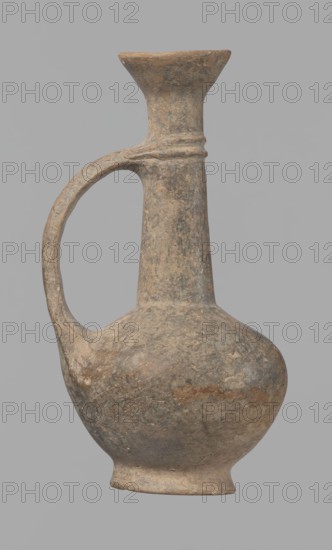 Jug, Cypriot pottery, Base Ring Ware I. Jug with an echinus-shaped foot; a biconical, slightly rounded belly; a high, narrow neck terminating in a funnel-shaped, flared spout. The neck is set at a slight inclination from the plane of the foot. A ribbon-shaped handle is mounted two-thirds up the neck and to the body; at the neck it merges into a double applied ring surrounding the neck. Pinkish clay with a brown surface, in places fading to a brick-red hue. Surface heavily rubbed, slight loss of the rings on the neck. 1600–1450 BC, Cyprus; vessel; height 12.3 cm, diameter 6.7 cm., An aged ceramic vessel with a rounded body, a narrow neck that widens into a flared rim, and an arched handle connecting the neck to the shoulder; the surface shows a worn, textured appearance with patches and speckles. The visible colors include muted browns, grayish tones, and subtle beige areas, with some darker mottling and traces of lighter, almost pale spots against a neutral gray background.