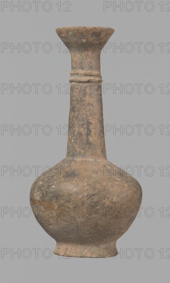 Jug, Cypriot pottery, Base Ring Ware I, 1600–1450 BC. Jug with an echinus-shaped foot; a biconical, slightly rounded belly; a high narrow neck terminating in a funnel-shaped, flared spout. The neck is set at a slight inclination from the plane of the foot. Ribbon-shaped handle mounted two-thirds up the neck and to the vessel body; at the neck it merges into a double applied ring surrounding the neck. Pinkish clay with a brown surface, locally fading to a brick-red hue. Surface heavily rubbed, slight loss of the rings on the neck. Origin: Cyprus Nea Paphos excavations. Vessel; height 12.3 cm, diameter 6.7 cm., A worn, weathered clay vessel with a rounded bulbous body tapering into a long narrow neck and flared rim, showing a rough textured surface with patches of muted brown, tan, gray, and darker sooty areas, with a small raised ring detail on the neck and an uneven base.