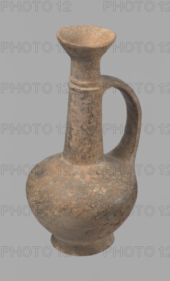 Jug, Cypriot pottery, Base Ring Ware I, 1600–1450 BC, Cyprus. Jug with an echinus-shaped foot; a biconical, slightly rounded belly; a high narrow neck terminating in a funnel-shaped, flared spout. The neck is set at a slight inclination from the plane of the foot. Ribbon-shaped handle mounted two-thirds up the neck and to the body; at the neck it merges into a double applied ring surrounding the neck. Pinkish clay with a brown surface, in places fading to a brick-red hue. Surface heavily rubbed with slight loss of the rings on the neck. Vessel, height 12.3 cm, diameter 6.7 cm., A single worn clay vessel with a rounded body, a narrow tall neck that flares into a funnel-shaped rim, and a curved handle connecting the neck to the shoulder; the surface shows uneven texture and pitting with areas of discoloration. The visible colors include shades of brown, tan, beige, and gray with darker mottled patches. The background is a uniform light gray.