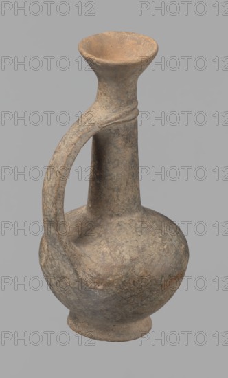 Jug. Cypriot pottery, Base Ring Ware I. Jug with an echinus-shaped foot; a biconical, slightly rounded belly; a high narrow neck terminating in a funnel-shaped, flared spout. The neck is set at a slight inclination from the plane of the foot. A ribbon-shaped handle is mounted two-thirds up the neck and to the arms of the vessel; at the neck it merges into a double plastic ring surrounding the neck. Pinkish clay with a brown surface, in places fading to a brick-red hue. The surface is heavily rubbed, with slight loss of the rings on the neck. 1600–1450 BC, Cyprus; vessel; height 12.3 cm, diameter 6.7 cm., An aged ceramic jug with a rounded body, a narrow tall neck that flares into a wide funnel-like mouth, and a single curved handle connecting the neck to the shoulder; the surface appears rough and weathered with mottled tones of gray, brown, tan, and hints of muted ochre, and the background is a flat light gray.