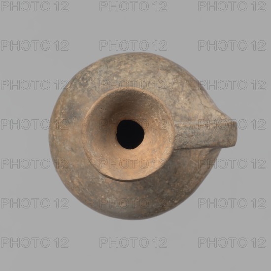 Jug, Cypriot pottery, Base Ring Ware I. Jug with an echinus-shaped foot; a biconical, slightly rounded belly; a high and narrow neck terminating in a funnel-shaped, flared spout. The neck is mounted at a slight inclination from the plane of the foot. Ribbon-shaped handle mounted two-thirds up the neck and to the arms of the vessel; at the neck it merges into a double plastic ring surrounding the neck. Pinkish clay, brown surface, in places fading to a brick-red hue. Surface heavily rubbed, slight loss of the rings on the neck. 1600–1450 BC, Cyprus. Vessel; height 12.3 cm, diameter 6.7 cm., A round vessel with a short neck and a funnel-shaped opening viewed from above, showing a smooth rim and a dark interior, with a single handle extending from the body; the surface appears worn and textured with mottled tones of brown, tan, gray, and hints of ochre against a neutral gray background.