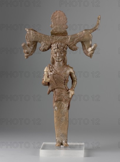 Mirror support with a depiction of the goddess Aphrodite. Support for a standing mirror; hand mirror; figurine of Aphrodite wearing an Ionian chiton and himation. Greek deity depiction, ca. 540–550 BC attributions: Zdrojewska places it on Samos ca. 540 BC; Franck and Furtwängler on the Peloponnese or in Arcadia 550–530 BC; Wallenstein in Corinth, dated to 550 BC. Samos place of origin. Mirror, height 23.2 cm, width 14 cm, depth 3 cm. Maker, inscription/author unknown., A standing bronze figurine with a brownish-green patina mounted on a clear rectangular base, depicting a humanlike figure wearing a long skirt and adorned with a large horizontal headdress that extends outward on both sides and ends in animallike heads; the figure has stylized facial features and textured hair, one hand resting near the hip while the other holds a small object, the surface shows cracks and uneven texture, and the background is a smooth gray.