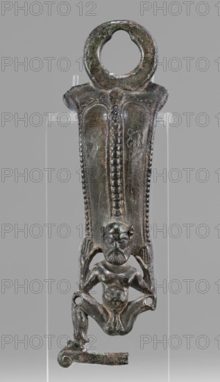Situla handle with a depiction of Silenus; unknown creator; Silenus mythology; bronze vessel; 1st half of the 5th century BC; Etruria; vessel; height 13.7 cm; width 4.2 cm, The image shows a vertical metal object with a patinated surface in shades of green, brown, and dark gray, mounted against a neutral gray background. The top of the object is rounded with a circular opening and a textured rim. The central section tapers downward and features a raised vertical ridge decorated with a row of bead-like protrusions and bordered by parallel dotted edges. Below this, a small sculpted humanoid figure is integrated into the lower portion, depicted with a bearded face, distinct facial features, a bare torso, and bent legs forming a squatting posture with feet and lower limbs extending outward. The figure appears attached to the broader upper section by its raised arms and sides, and the lower edge of the object includes a short horizontal extension with a curved end. The object rests on a tra...