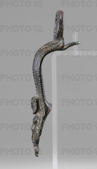 Situla handle with a depiction of Silenus; maker, Silenus mythology; bronze vessel; 1st half of the 5th century BC; Etruria; vessel; height 13.7 cm; width 4.2 cm., A vertical metal object with a curved upper loop and an elongated S-shaped body is shown against a plain gray background; the object has a dark metallic surface with areas of greenish patina and brownish discoloration, a beaded raised ridge running along the outer curve, and a small sculpted human-like figure attached to the lower portion with a distinct head, facial features, and a torso; the piece rests on a translucent clear support with a few clear pegs visible.